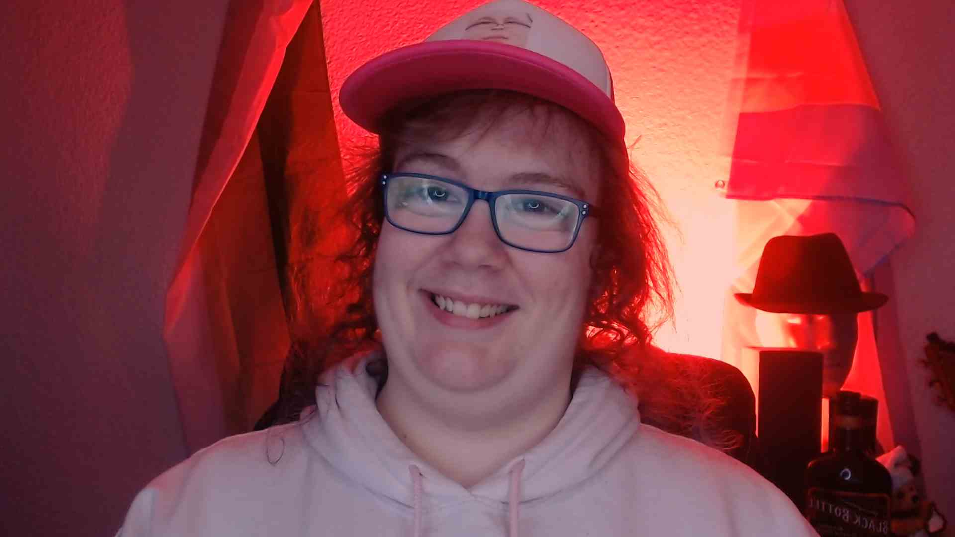 A picture of me looking straight at the camera with a big smile. I am wearing a pink hoodie, a fuschia and white hat which also has a picture of me on it. The background is lit with a bright red light
